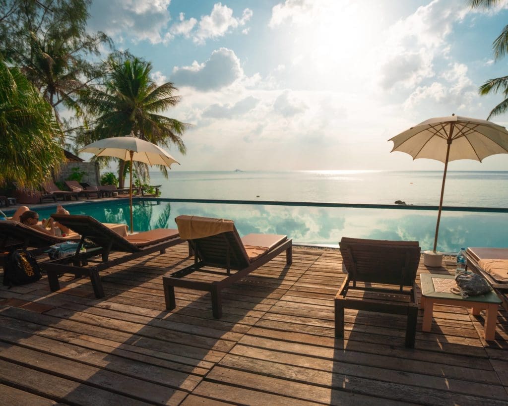 Lounge chairs and parasols by an infinity pool at luxury resorts overlook a calm sea under a partly cloudy sky at sunrise or sunset.