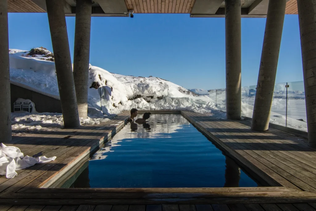 Two people relax in a rectangular outdoor pool under a wooden roof, surrounded by snow-covered landscape and concrete pillars.