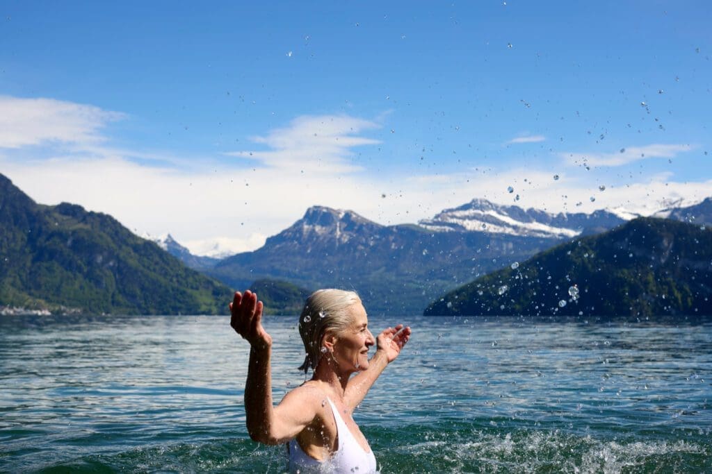 An older woman in a white swimming costume stands in a lake with arms raised, surrounded by mountains and a blue sky—embracing the serenity often sought through luxury services.