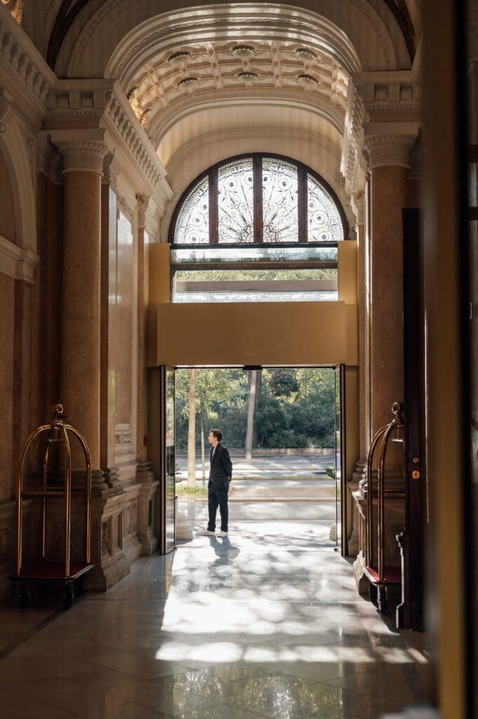 A person in a suit stands near the open glass doors of an ornate building lobby with marble columns and sunlight streaming in.