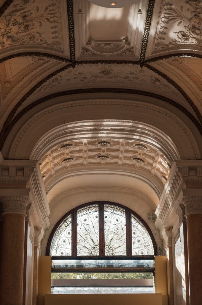 Ornate arched ceiling with decorative plasterwork above a large round window featuring intricate ironwork and yellow architectural elements below.