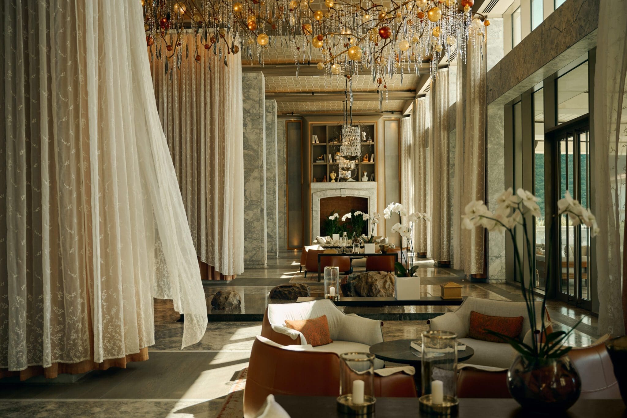 Elegant hotel lounge with marble floors, sheer curtains, modern seating, chandeliers, and large windows letting in natural light. Orchids and candles decorate the space.