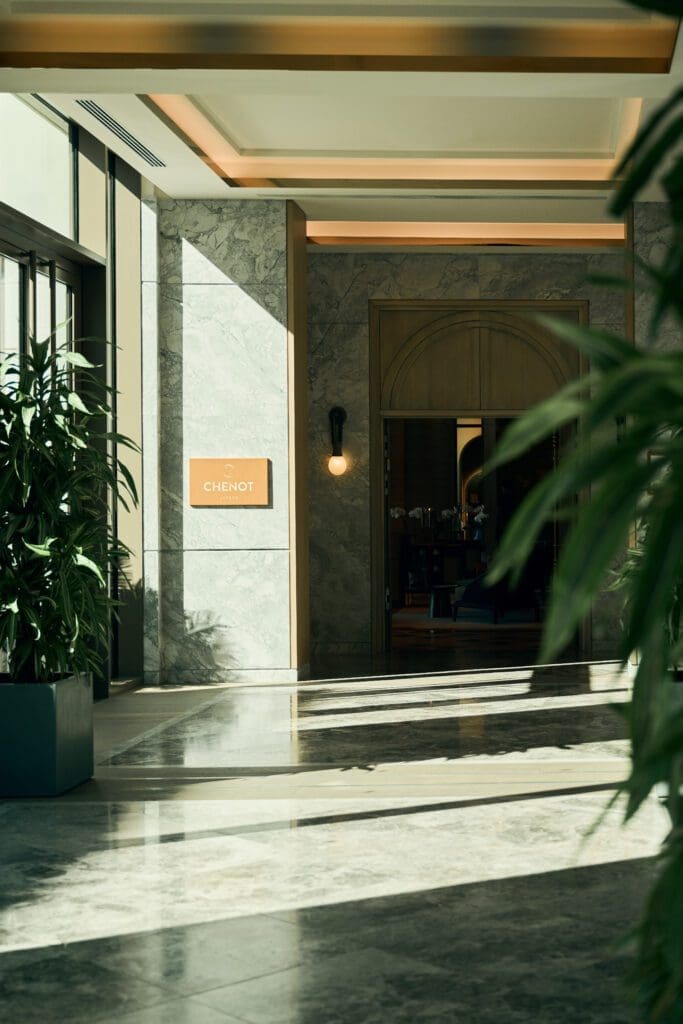 A sunlit corridor with marble floors and walls, potted plants, and a sign reading "CHENOT" next to an arched wooden doorway.