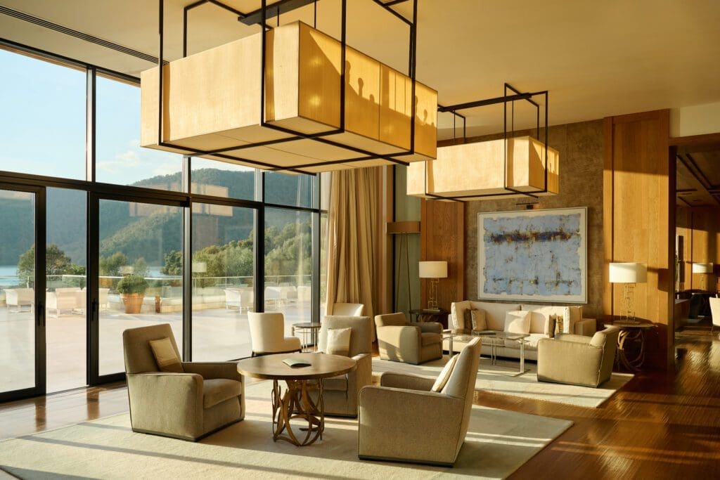 A modern lounge with beige armchairs, large square ceiling lights, wooden floors, floor-to-ceiling windows, and a view of outdoor greenery and mountains.