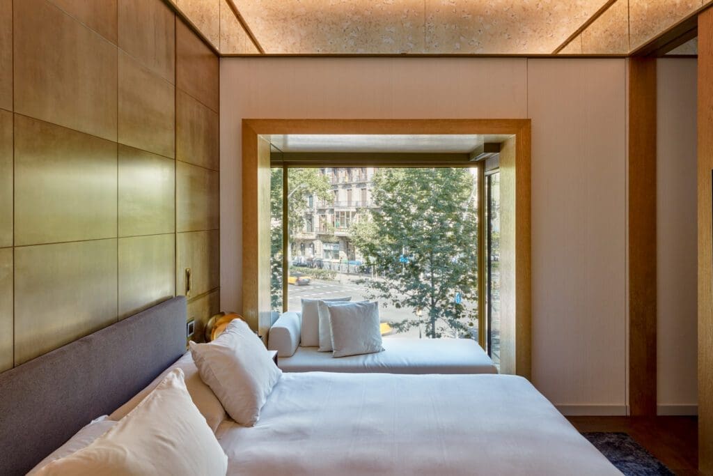 Modern hotel room with a neatly made bed, large window seat, and floor-to-ceiling window offering a view of trees and a city street outside.