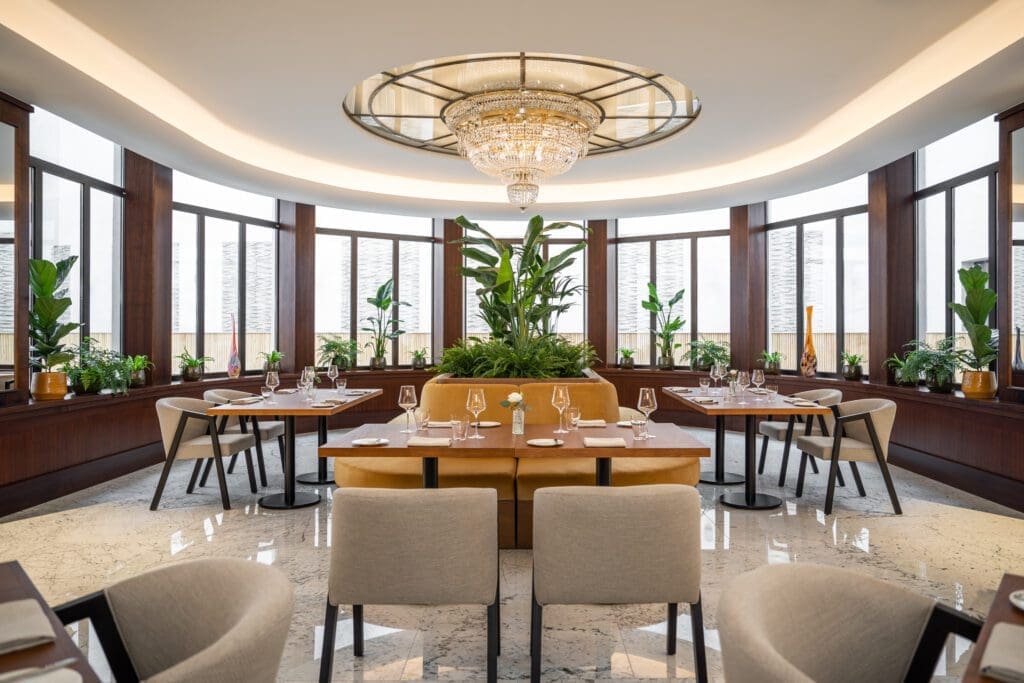 Modern restaurant dining area with round chandelier, large windows, green plants, beige chairs, and set tables on polished marble floor.