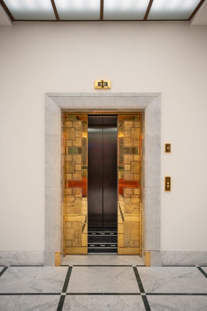 An open lift with gold and marble trim is centred in a white corridor with marble floor and walls; buttons and an indicator light are visible beside the door.
