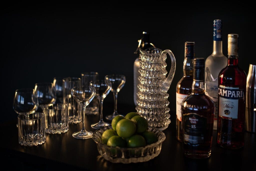 A selection of spirit bottles, glasses, a glass jug, and a bowl of limes are arranged on a dark surface against a dark background.