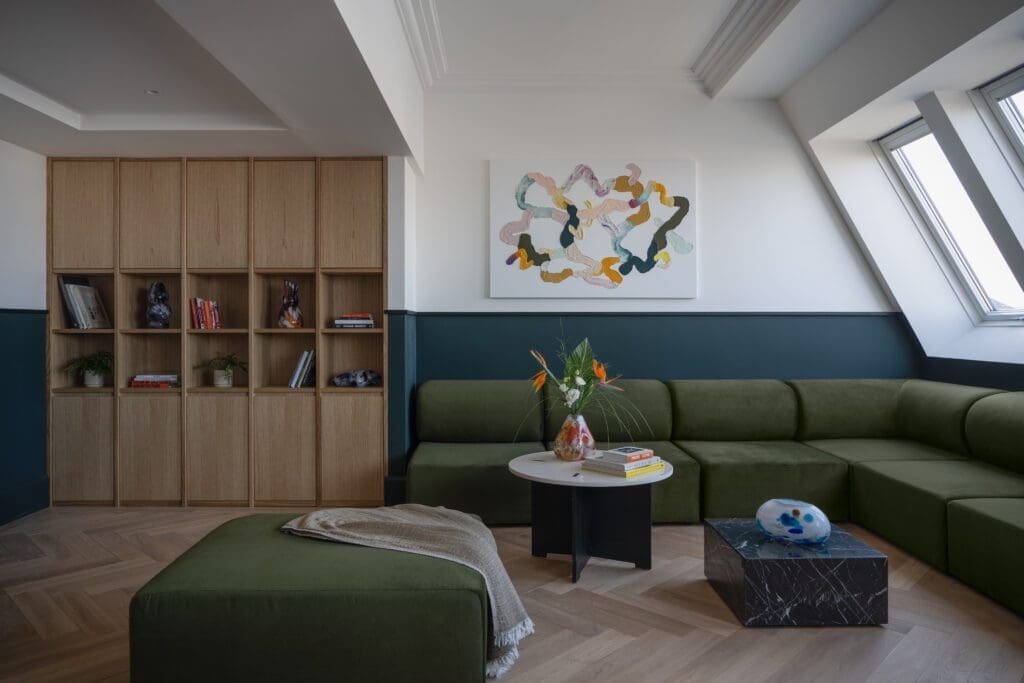 A modern living room with a green corner sofa, wooden bookcase, abstract wall art, and two coffee tables with books and a vase of flowers near large windows.