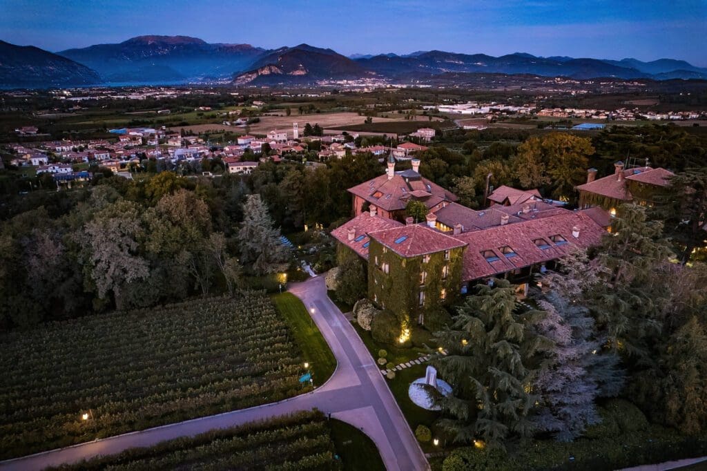 Aerial view of a large villa surrounded by trees and vineyards at dusk, with a town and mountains visible in the background.