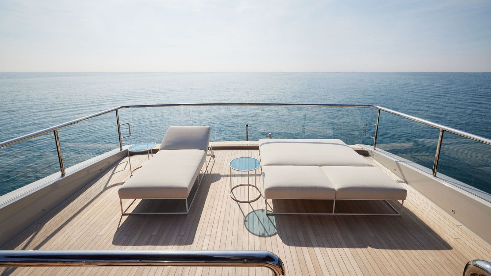 Two modern lounge chairs and small tables are arranged on a yacht deck overlooking a calm, open sea under a clear sky.