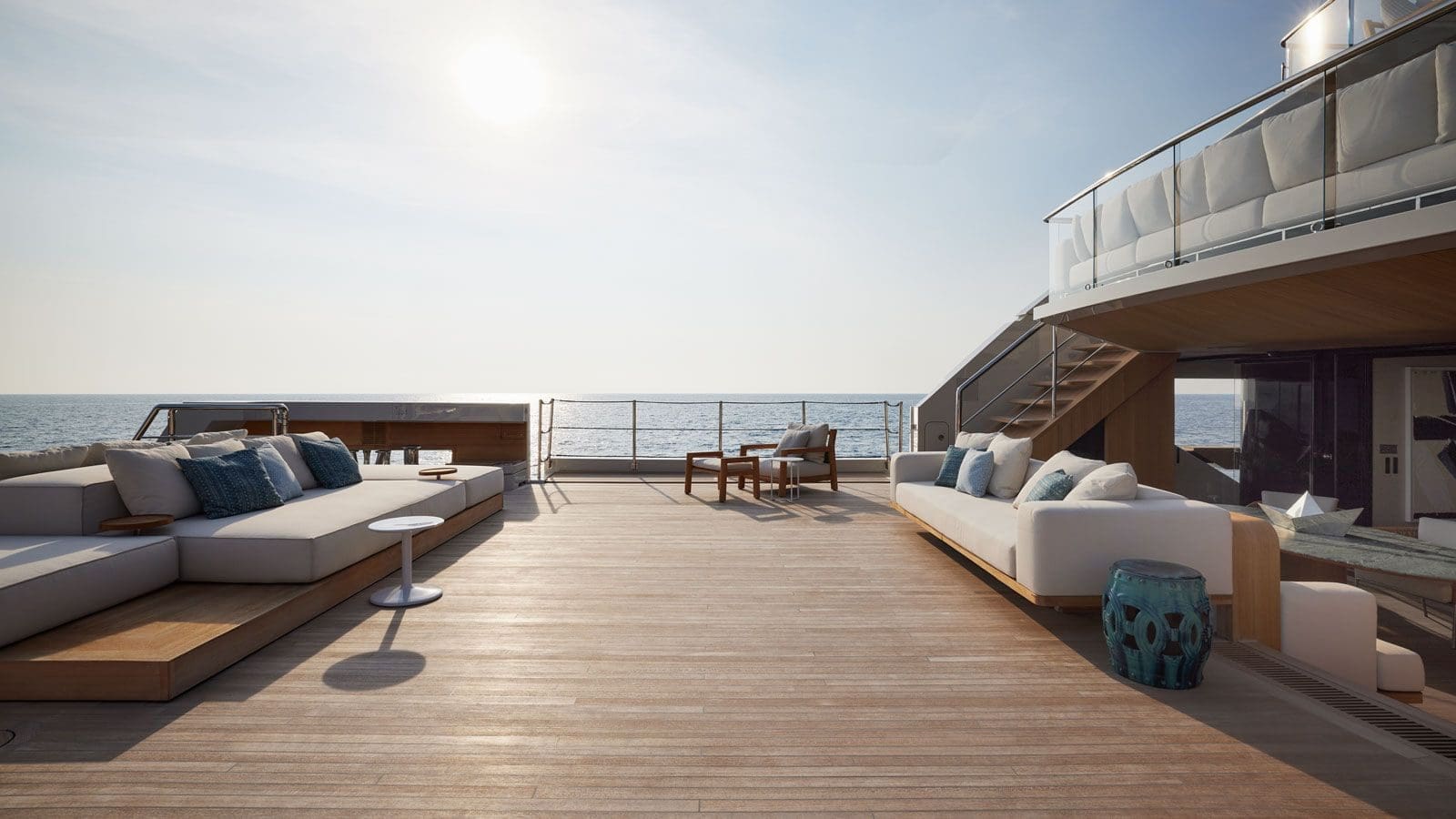 Spacious yacht deck with outdoor sofas, chairs, side tables, and sea view under a clear sky. Sun is low, casting soft light across the wooden flooring.