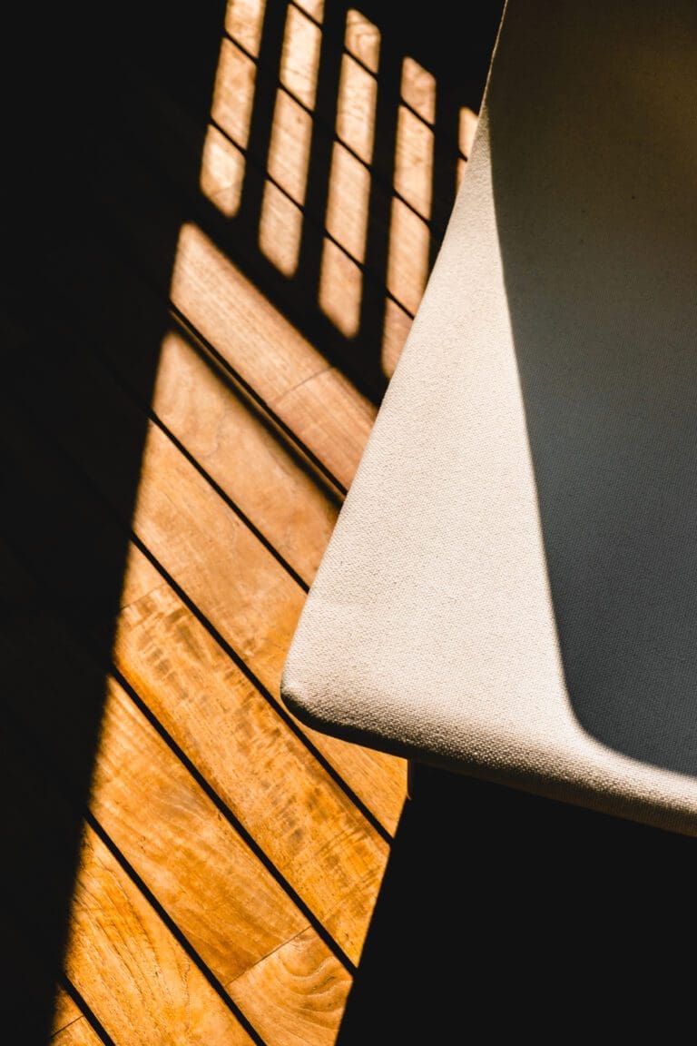 Sunlight casts shadows from railing onto wooden floor next to the edge of a light-coloured fabric chair.