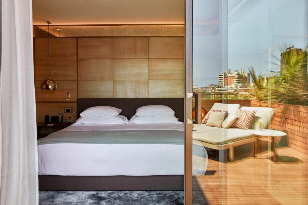 A modern hotel room with a neatly made bed, wood-panelled walls, and a glass door opening to a balcony with a lounge chair and city view.