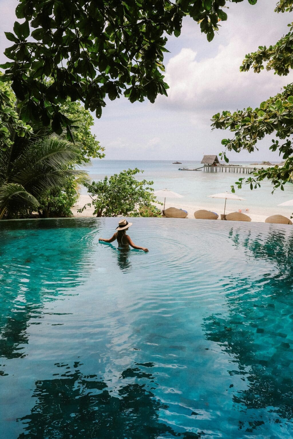 A person in a sunhat relaxes at the edge of an infinity pool overlooking a tropical beach and sea, with a pier and thatched hut visible in the distance.