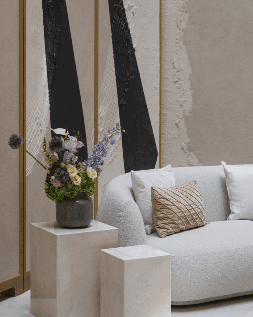 A modern living room with a white curved sofa, beige textured cushions, a vase of flowers on a square plinth, and abstract black and white wall art in the background.