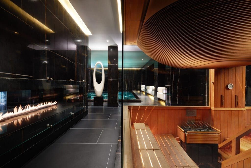 A modern spa interior with a wooden sauna on the right, a glass barrier, a long gas fireplace, and a sleek, dark-tiled relaxation area with ambient lighting.