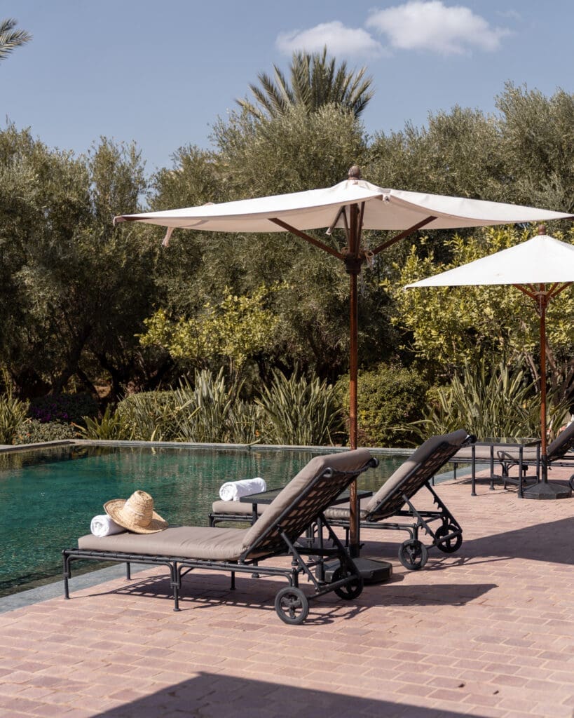 Poolside loungers with white umbrellas and rolled towels beside a clear swimming pool, surrounded by green trees and plants under a partly cloudy sky.