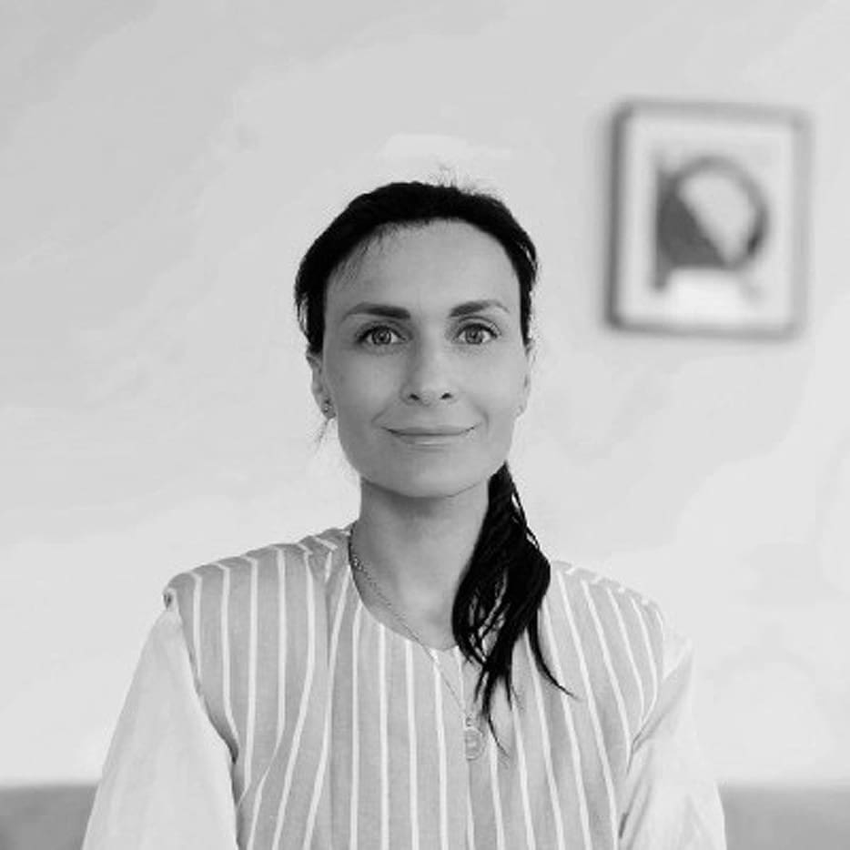A woman with dark hair pulled to one side is wearing a striped top and sits in front of a blank wall with a framed picture in the background. The photo is in black and white.