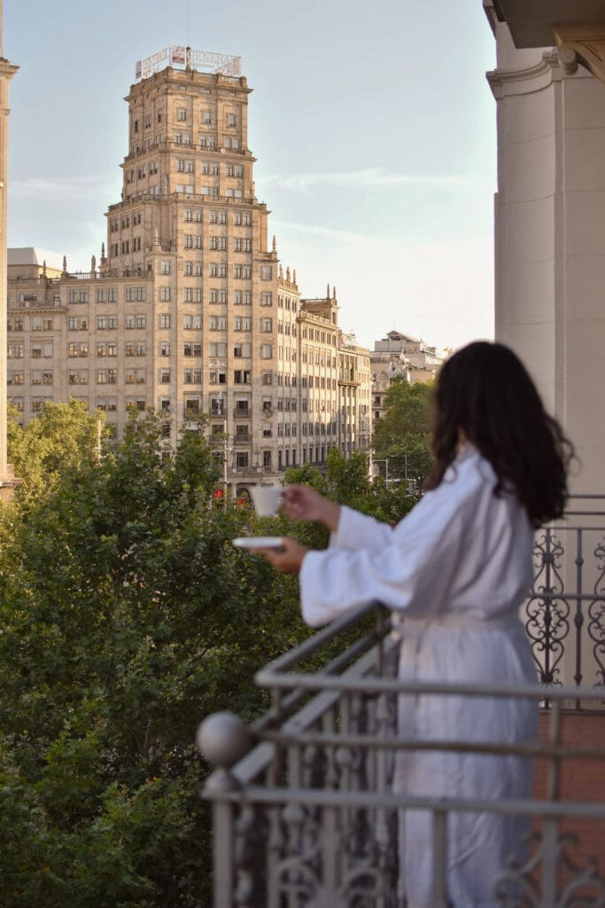 A person in a white dressing gown stands on a balcony holding a cup and saucer, overlooking a cityscape with a tall historic building and trees.