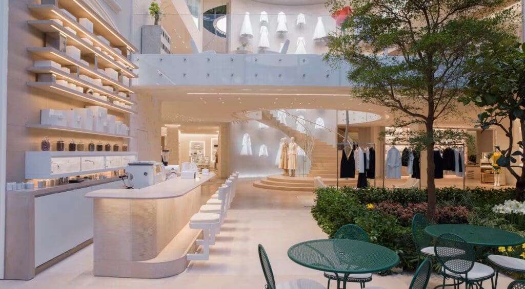 A modern luxury retail store with a minimalist café, spiral staircase, and curated clothing displays evokes desire and emotion. Green patio furniture and indoor plants complement the neutral-toned design in the inviting foreground.